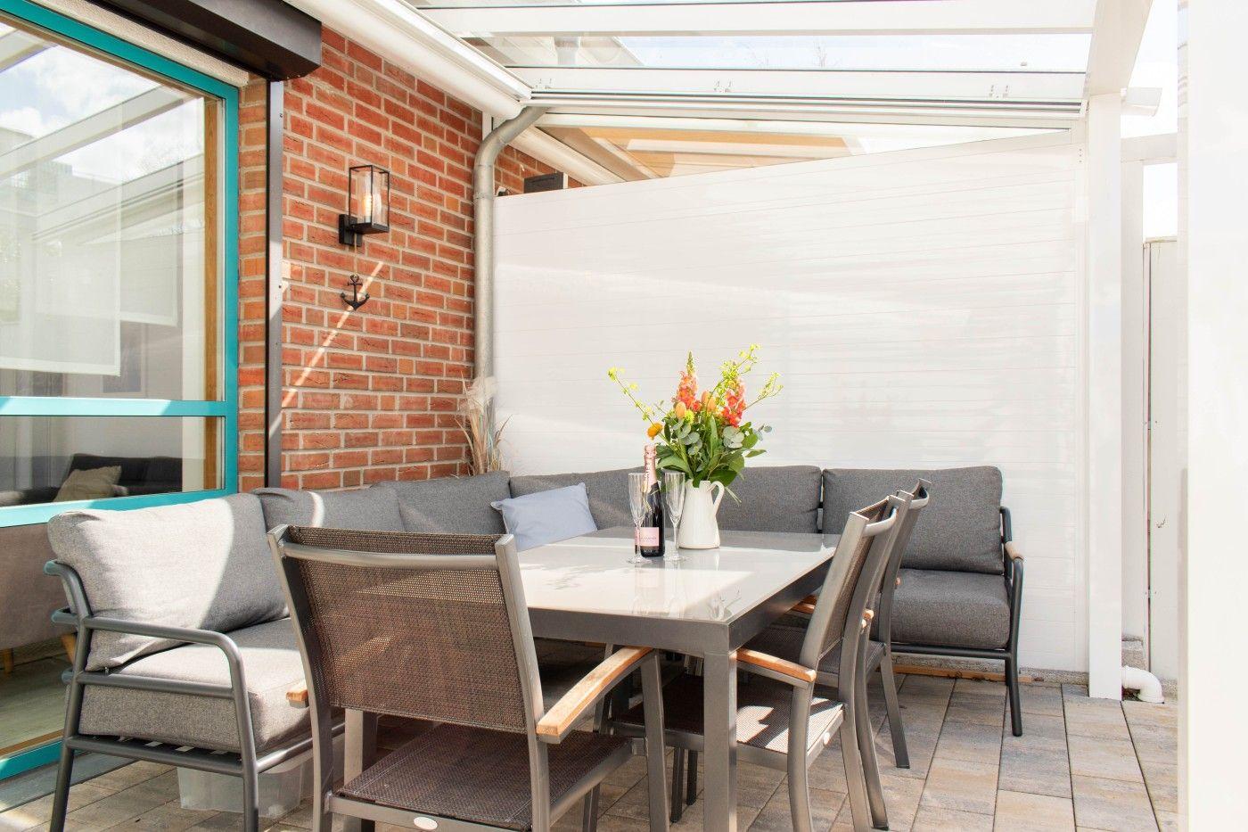 Terrasse mit Sitzgruppe, Tisch und Blumenvase unter Glasdach.
