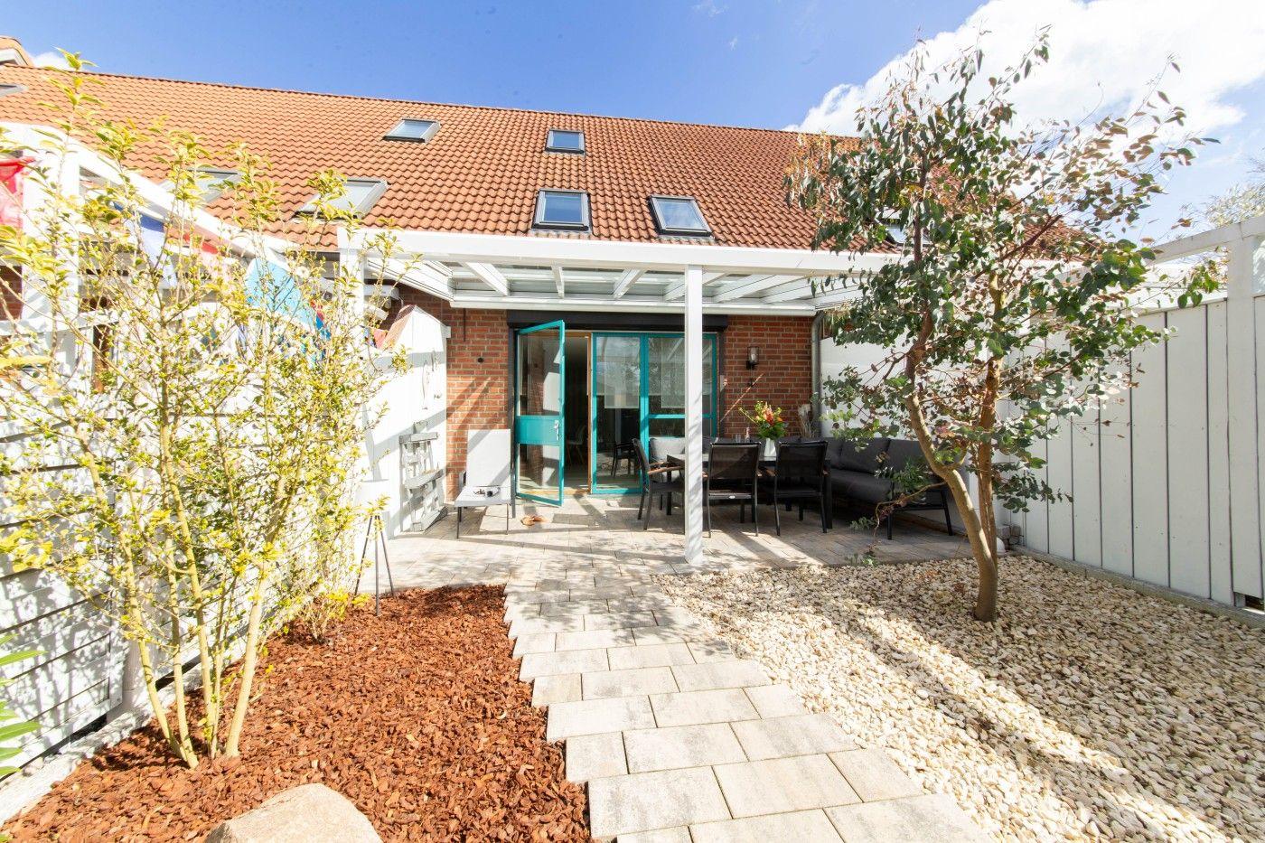 Hinterhof mit Terrasse, Sitzgelegenheiten und gepflastertem Weg. Haus mit rotem Dach und Fenstern.