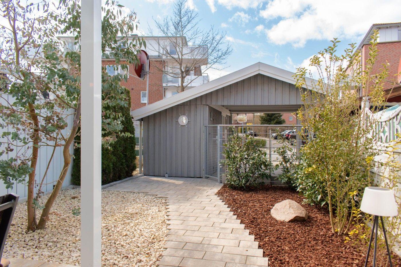 Gartenweg führt zu einer Garage mit Holzverkleidung und Metalltor. Umgeben von Pflanzen und Steinen.