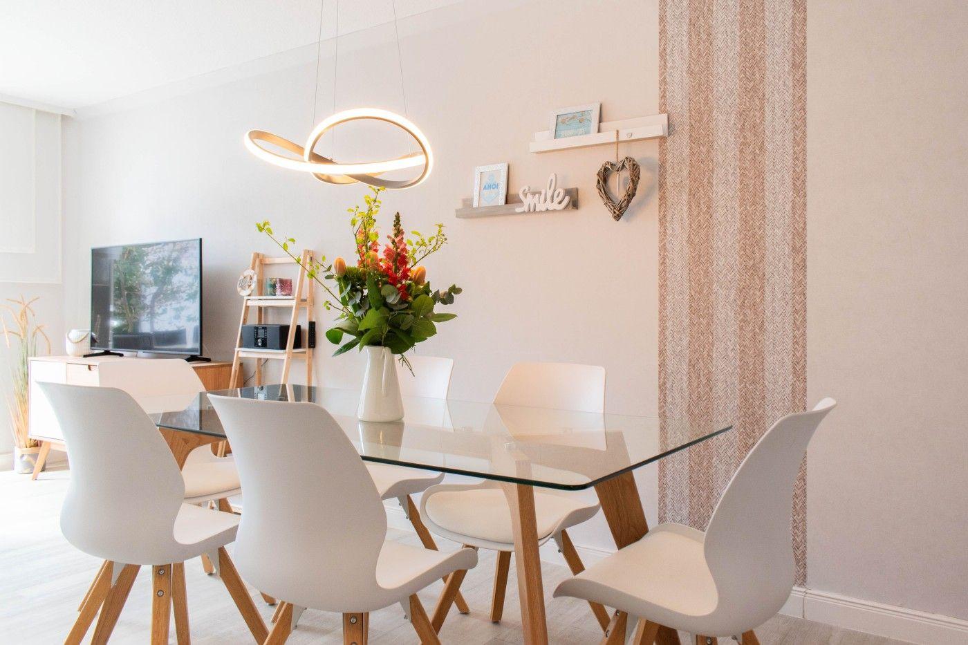 Esstisch mit Glasplatte und weißen Stühlen. Hintergrund: Wand mit Regal und Blumen.