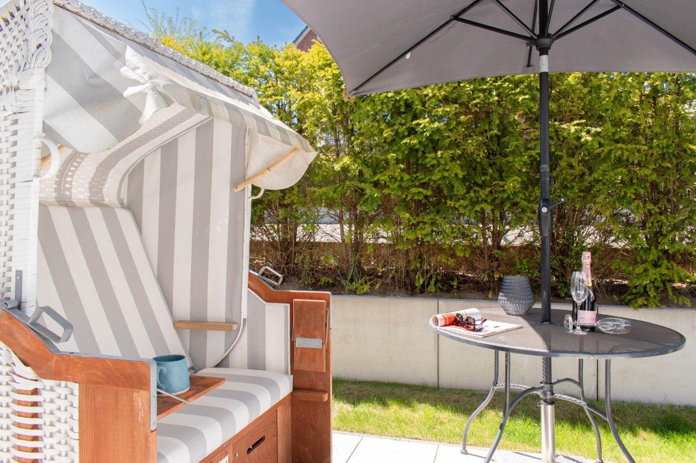 Terrasse mit Strandkorb, Tisch und Sonnenschirm. Hintergrund: grüne Hecke.
