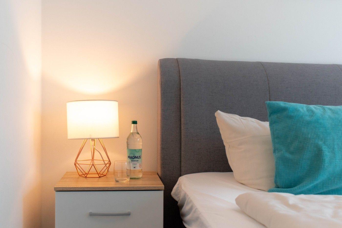 Schlafzimmer mit grauem Kopfteil, weißer Bettwäsche und blauem Kissen. Nachttisch mit Lampe und Flasche.