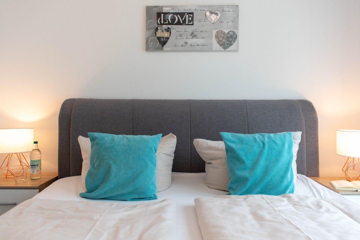 Doppelbett mit zwei türkisfarbenen Kissen und einem Wandbild mit Herzmotiven.