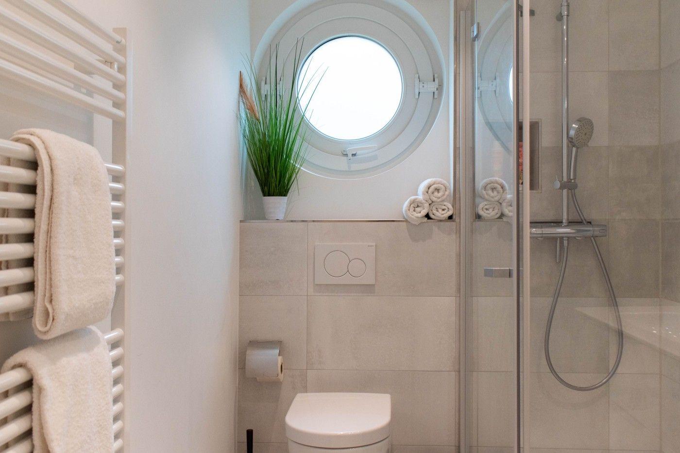 Badezimmer mit Dusche, Toilette und runden Fenster. Handtücher und Pflanze sind sichtbar.