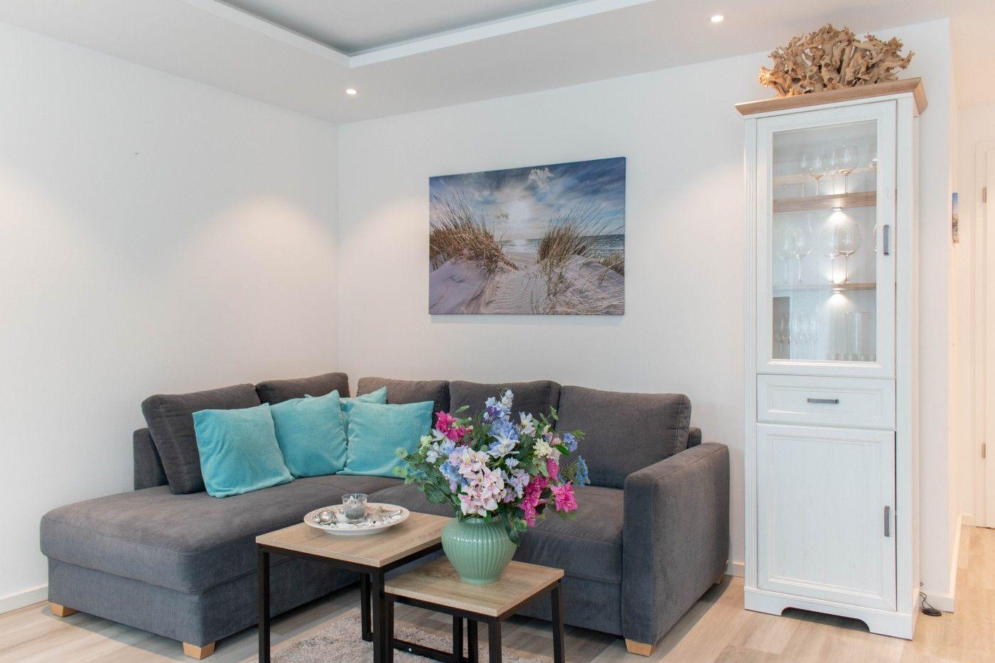 Ein Wohnzimmer mit grauem Sofa, blauen Kissen, Tisch mit Blumen und weißen Schrank.