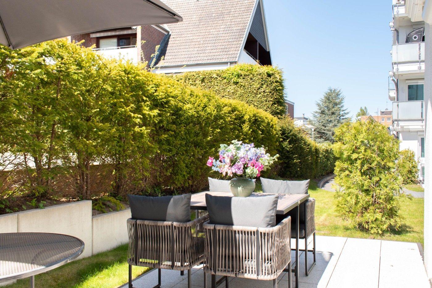 Terrasse mit Tisch, Stühlen und Blumenarrangement. Umgeben von grünen Hecken und Wohnhäusern.