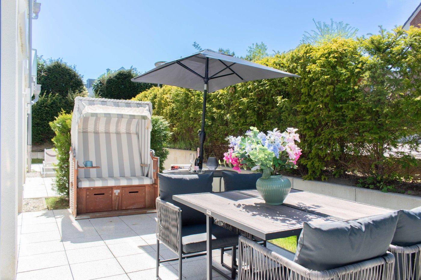 Terrasse mit Strandkorb, Tisch, Stühlen und Sonnenschirm. Blumen in Vase. Hintergrund: grüne Hecke.