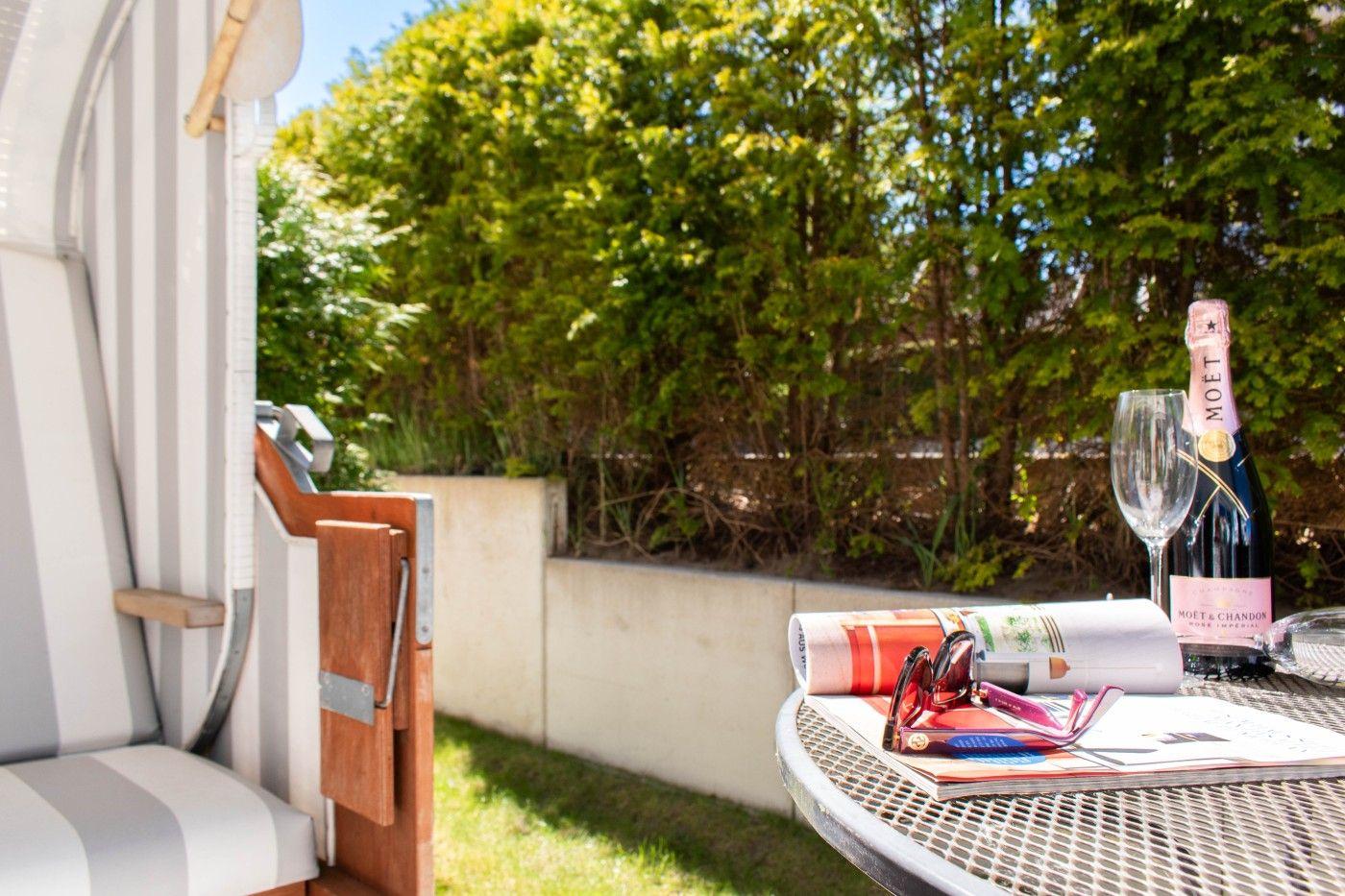 Außenbereich mit Tisch, Flasche Champagner und Sonnenbrille. Hintergrund: grüne Hecke.