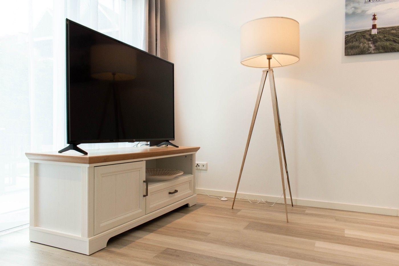 TV auf weißen Schrank mit Holzoberfläche, daneben Stehlampe und Wandbild.