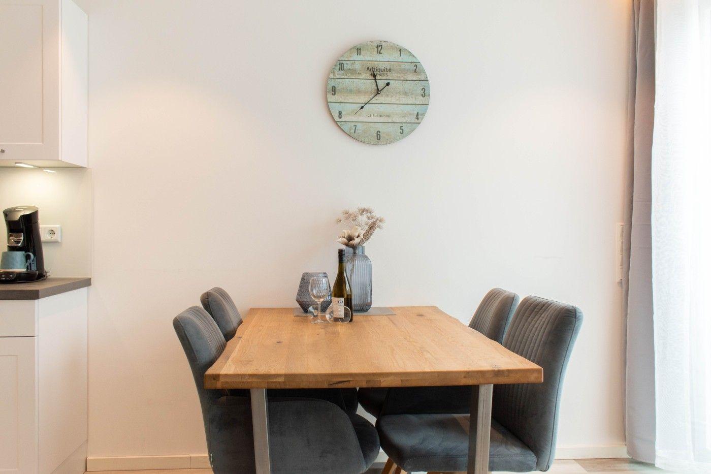 Esszimmer mit Holztisch, vier Stühlen und Wanduhr. Küchenzeile links, Fenster rechts.