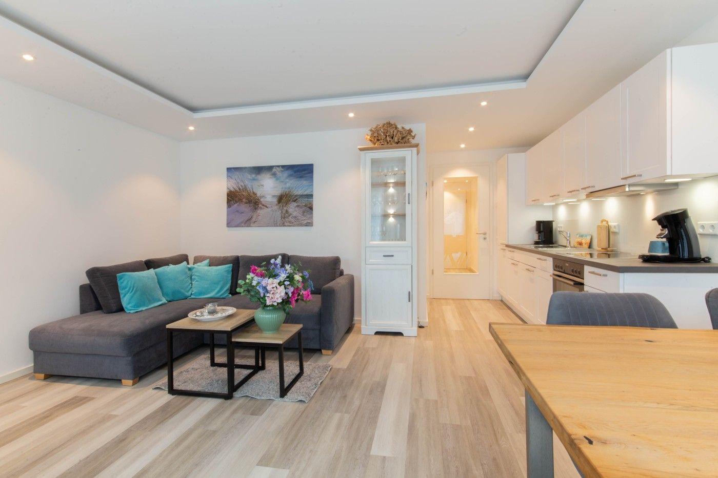Wohn- und Küchenbereich mit grauem Sofa, Holzboden und weißer Einbauküche.