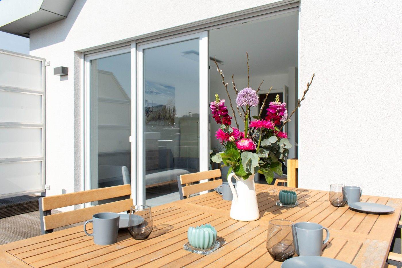 Terrasse mit Tisch, Stühlen und Blumenarrangement. Durchgehende Glasfront zum Innenraum.