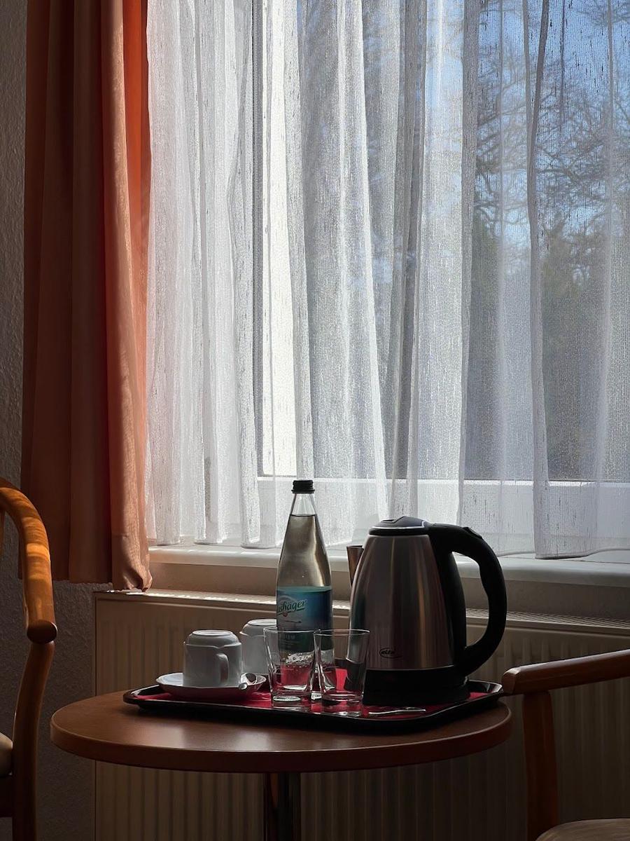Tisch mit Kaffeekanne, Gläsern und Flasche vor Fenster mit weißen Vorhängen.