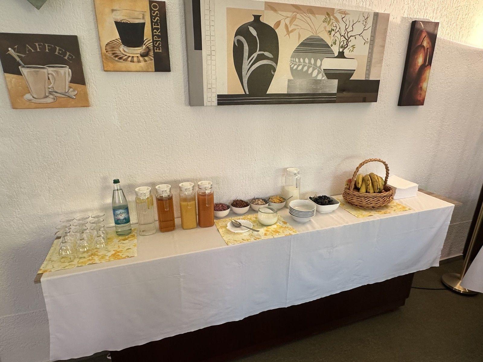 Frühstückstisch mit Getränken, Obst und Müslischalen vor Wand mit Bildern.