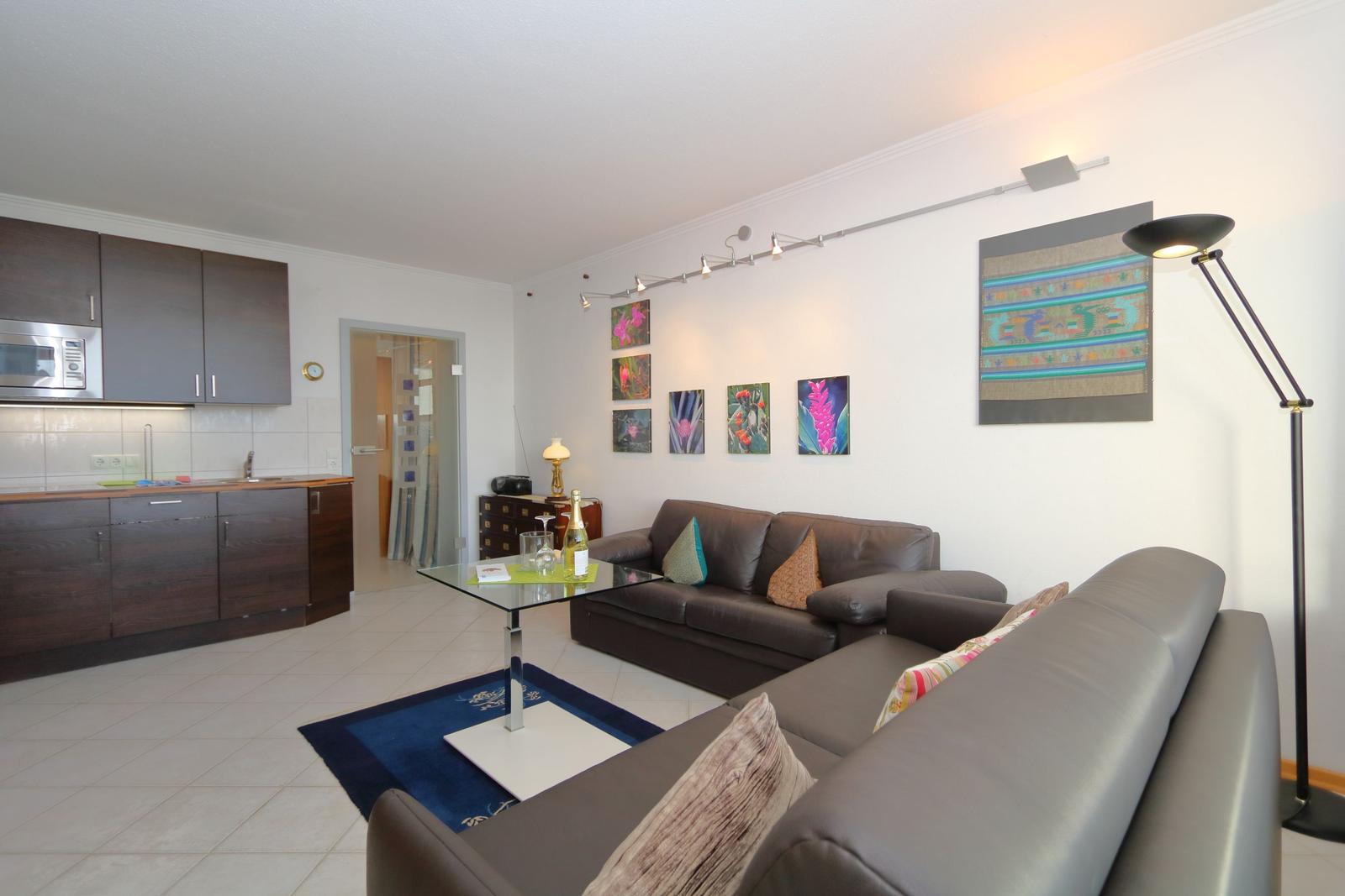Living room with sofa, coffee table, and kitchen area. Wall art and floor lamp visible.