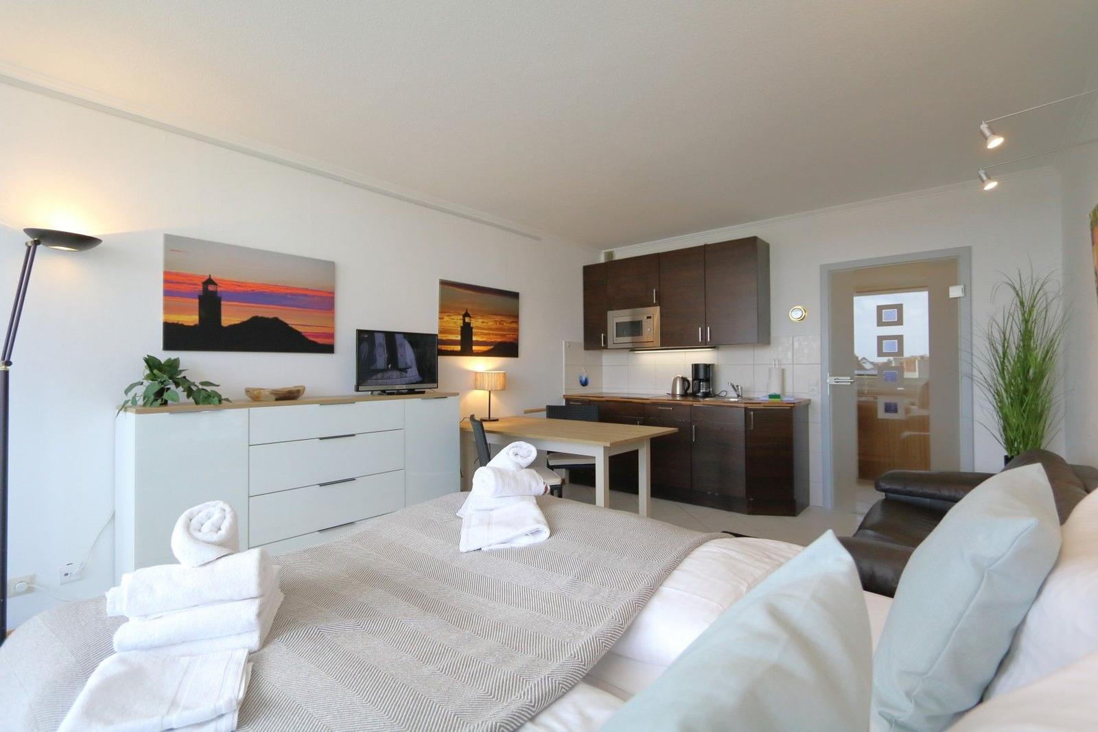 A bedroom with a bed, TV, and kitchen area in the background.