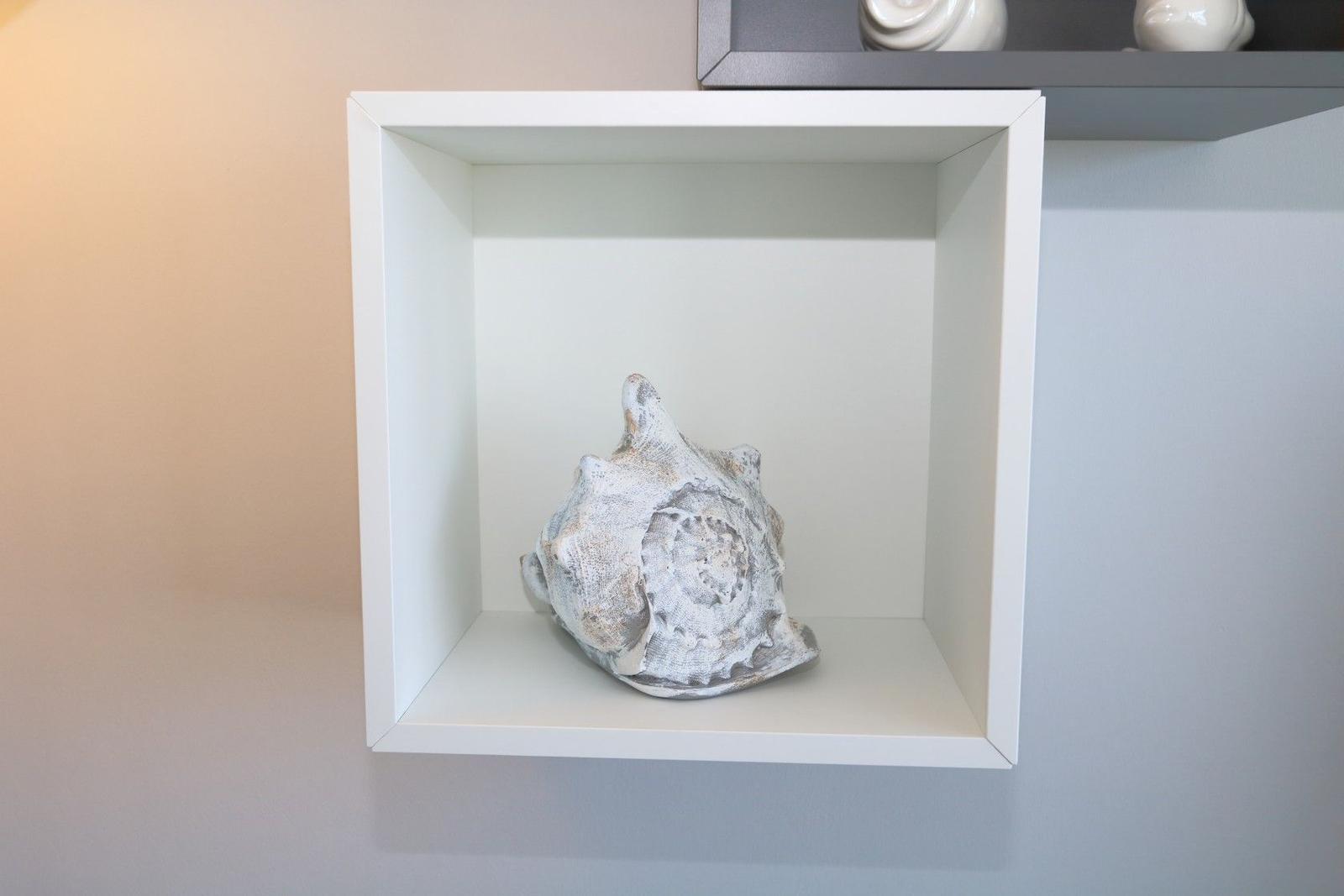 Eine weiße Wandnische mit einer Muschel. Oben sind weiße Objekte auf einem Regal.