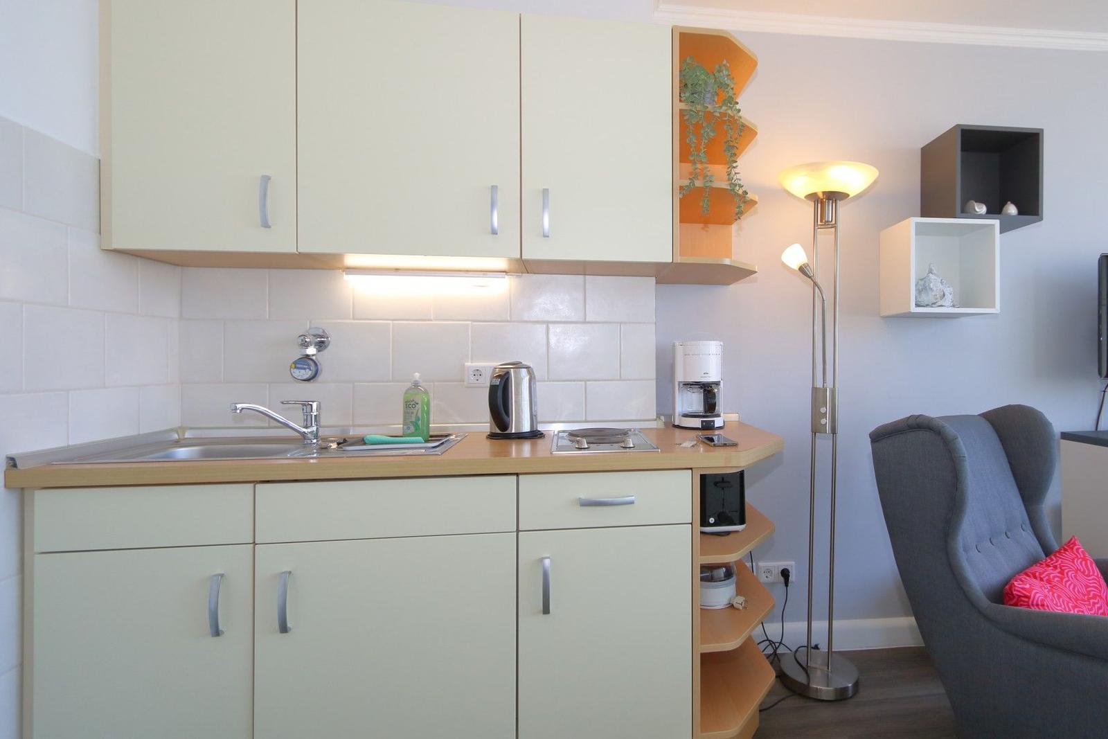 Küche mit Spüle, Kaffeemaschine und Stuhl. Wandregale und Stehlampe sind sichtbar.