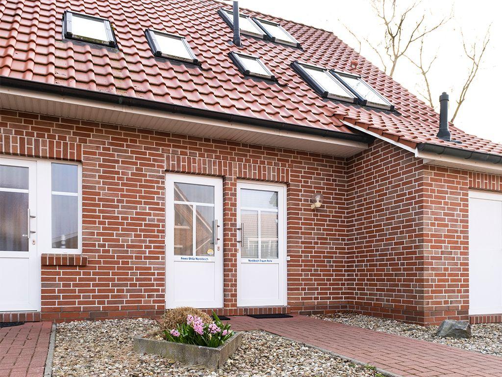 Ferienhaus mit rotem Ziegeldach und weißen Türen. Vorgarten mit Blumenbeet.