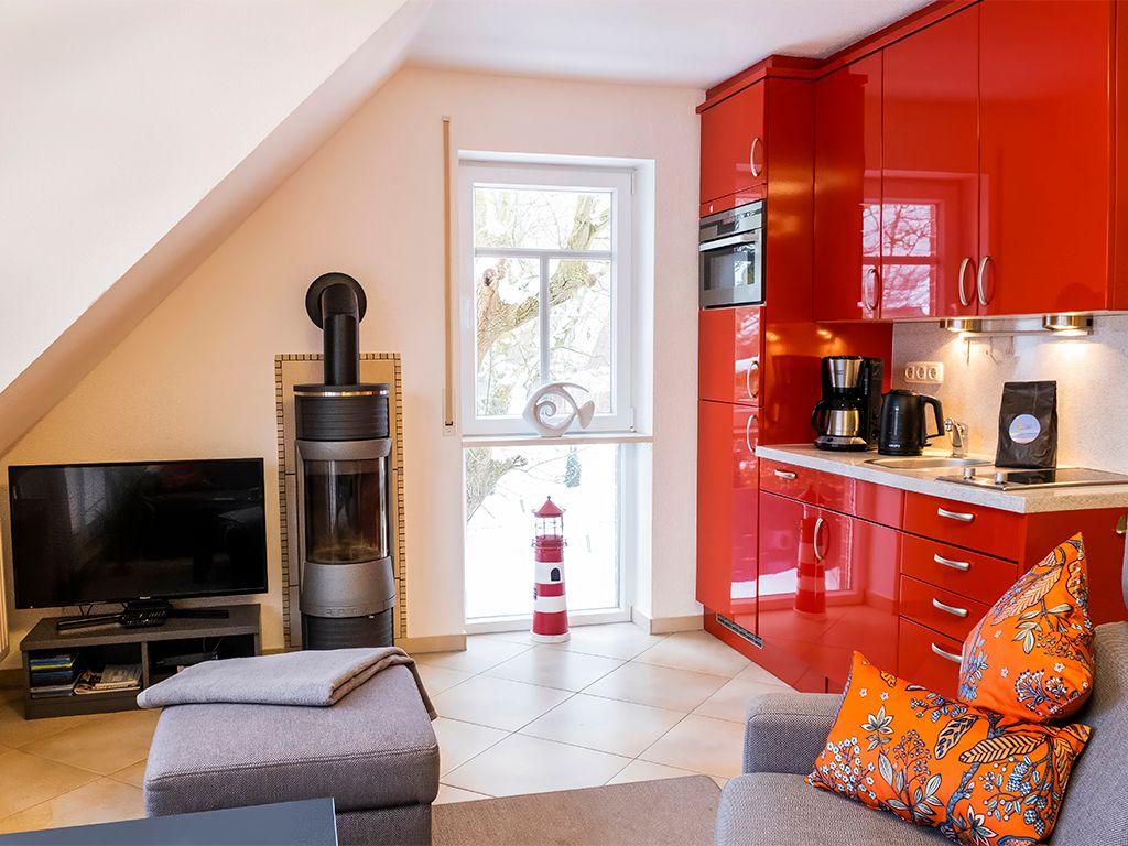 Wohnbereich mit roter Küche, Kaminofen und TV. Fenster mit Leuchtturm- Deko.