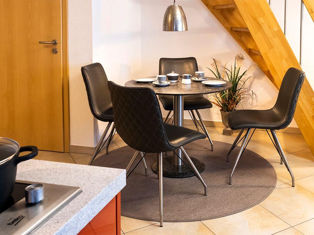 Runder Tisch mit vier schwarzen Stühlen unter einer Lampe. Holztreppe und Pflanze im Hintergrund.