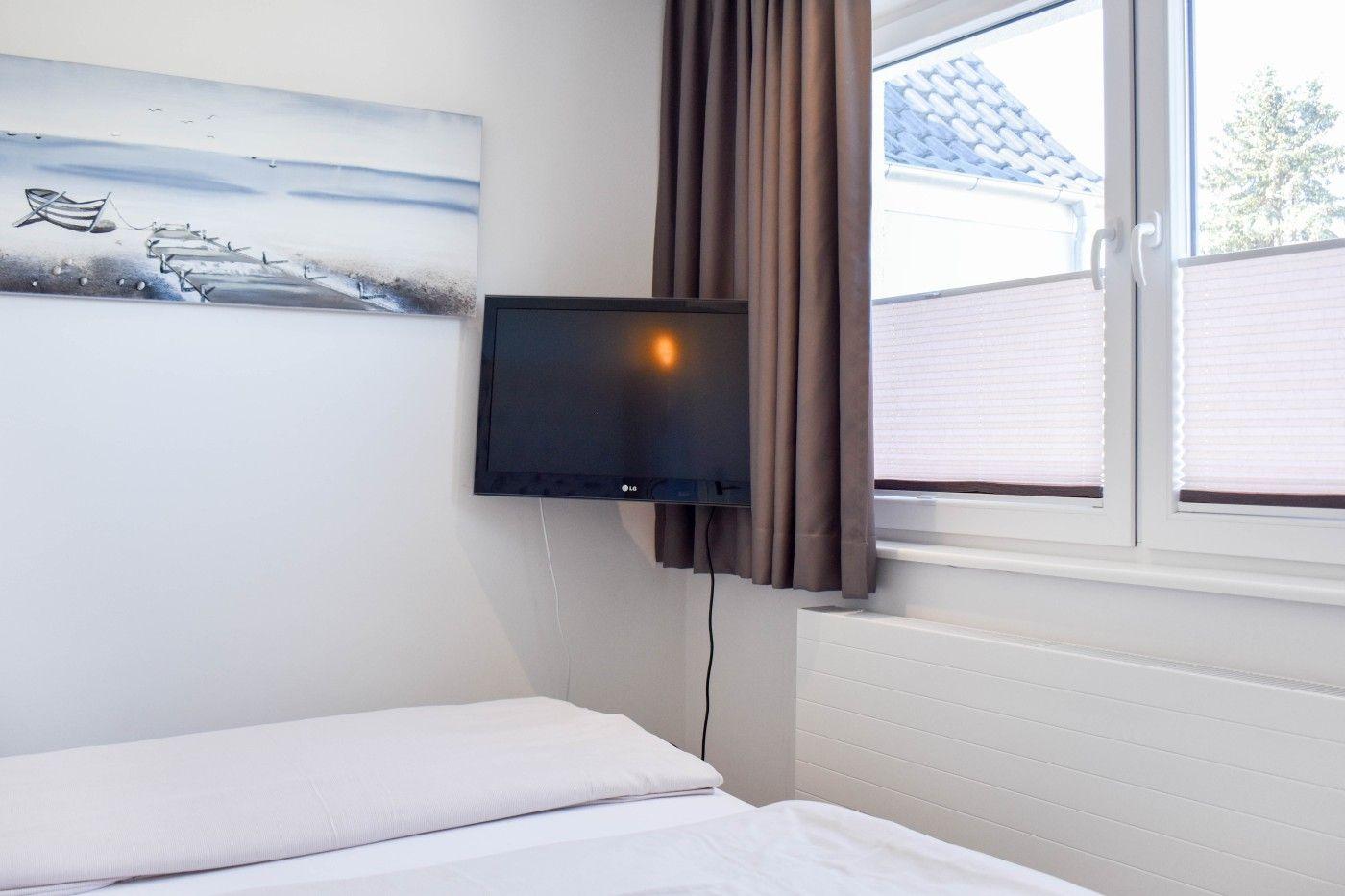 Schlafzimmer mit Bett, Wandbild und Fernseher an der Wand. Fenster mit Jalousie und Vorhang.