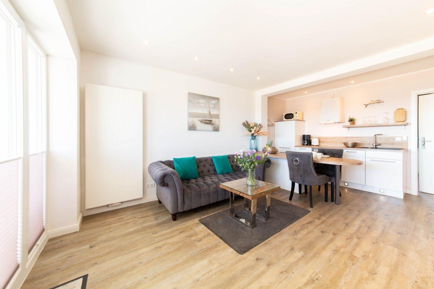 Wohnbereich mit Sofa, Esstisch und Küchenzeile. Holzboden und große Fenster.