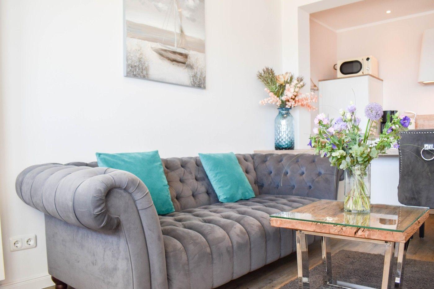 Ein grauer Polstersofa mit zwei türkisfarbenen Kissen und einem Glas-Tisch mit Blumen.