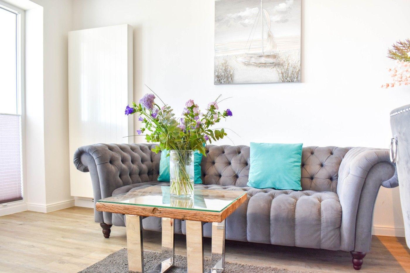 Ein grauer Chesterfield-Sofa mit türkisen Kissen und einem Glas-Tisch mit Blumen.