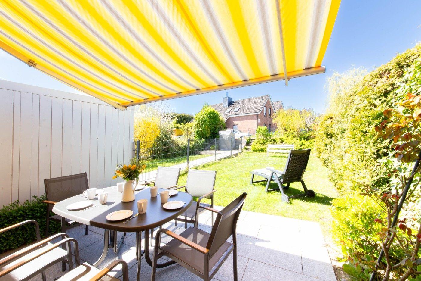 Terrasse mit gelb-weißem Sonnenschutz, Tisch, Stühle und Gartenblick.