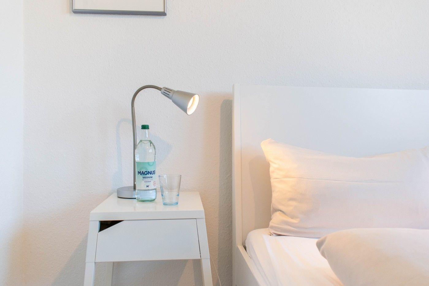 Nachttisch mit Lampe, Wasserflasche und Glas neben dem Bett.