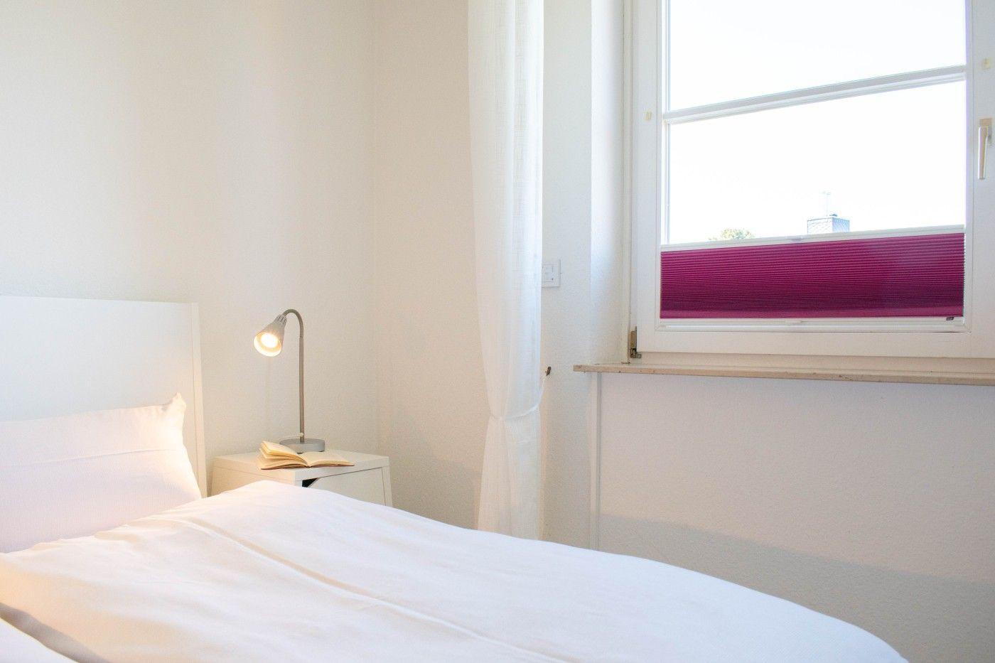 Schlafzimmer mit weißem Bett, Nachttisch und Fenster mit lila Jalousie.