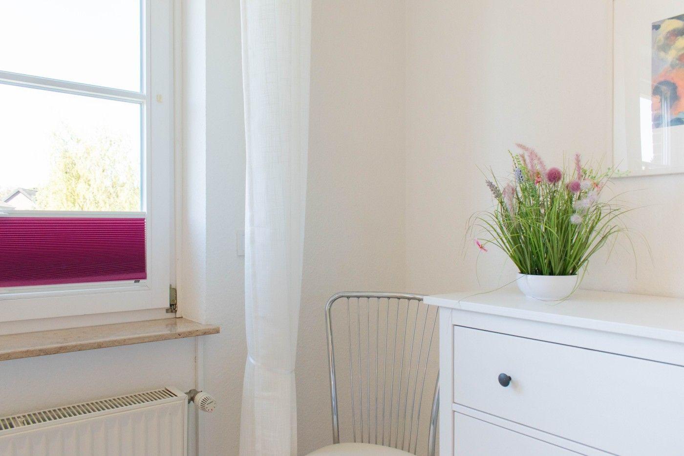 Ein Raum mit Fenster, weißem Schrank und Stuhl. Auf dem Schrank steht eine Blumenvase.