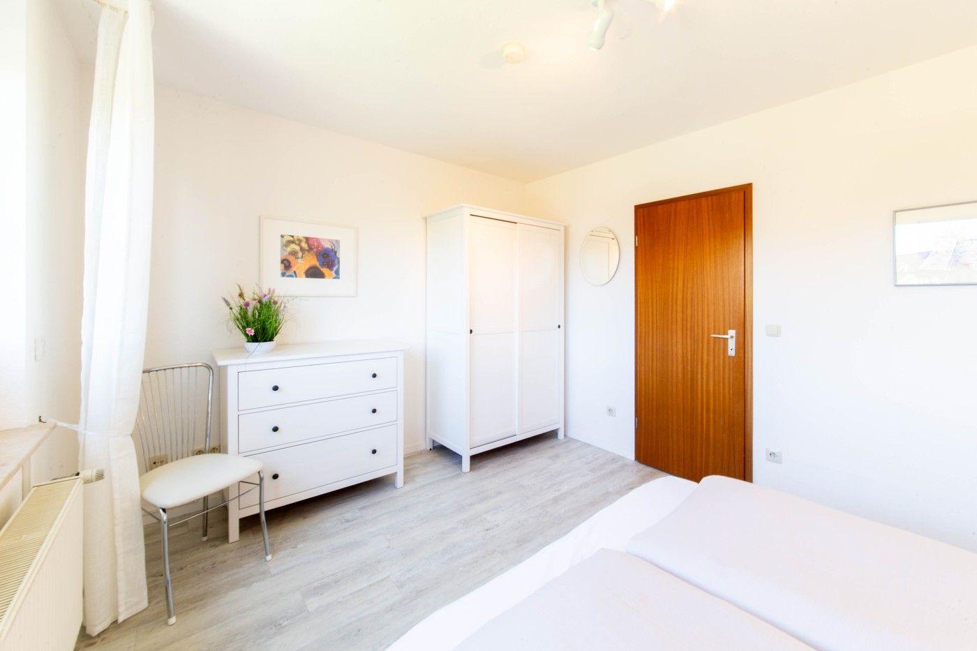 Schlafzimmer mit weißem Schrank, Schreibtisch und Bett. Holztür und Wandbilder sichtbar.
