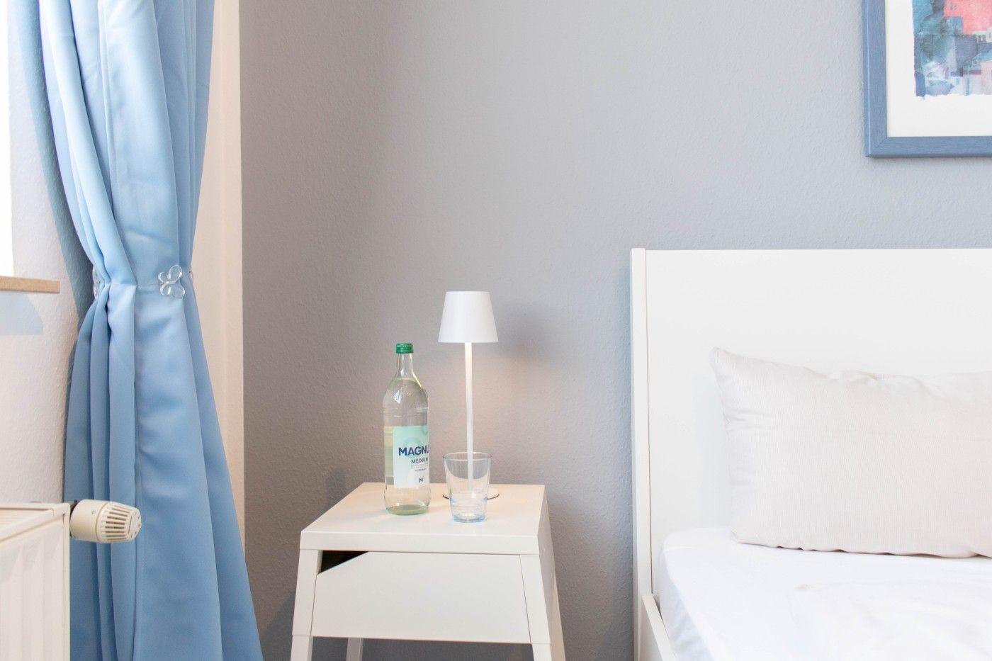 Schlafzimmer mit weißem Bett, Nachttisch, Lampe und Flasche.