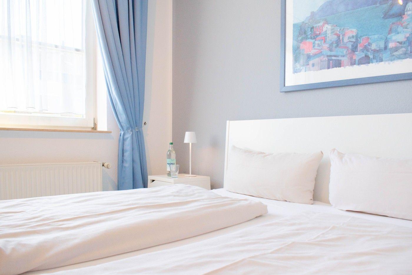 Schlafzimmer mit weißem Bett, blauen Vorhängen und Wandbild.