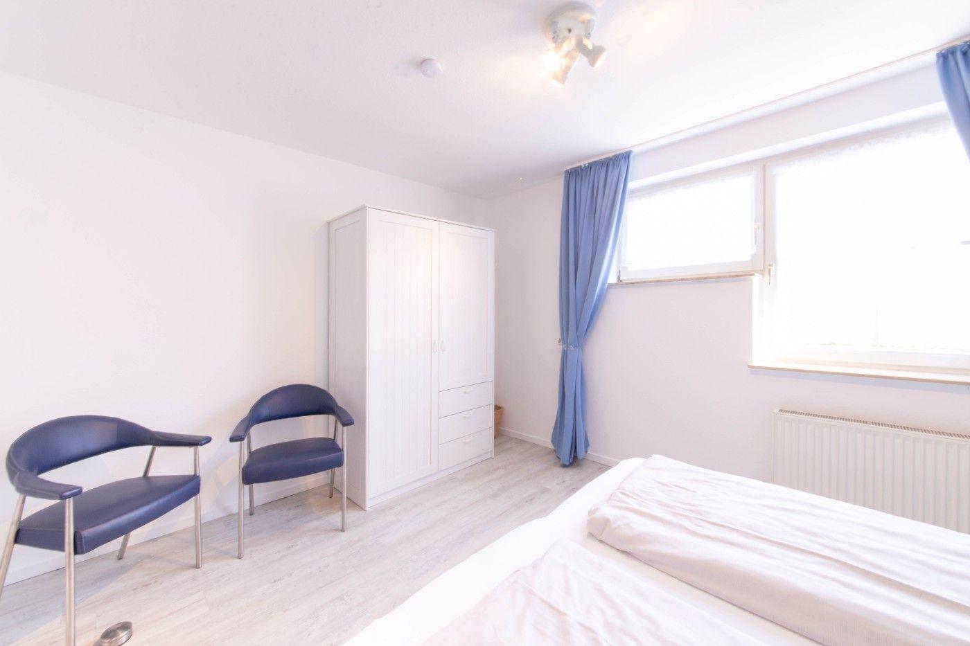 Schlafzimmer mit Bett, Kleiderschrank und zwei Stühlen. Fenster mit blauen Vorhängen.