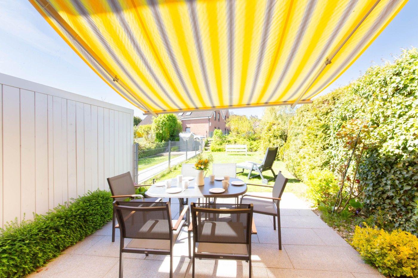 Terrasse mit Tisch, Stühlen und gelb-weißem Sonnensegel. Umgeben von grünen Hecken und einem weißen Zaun.