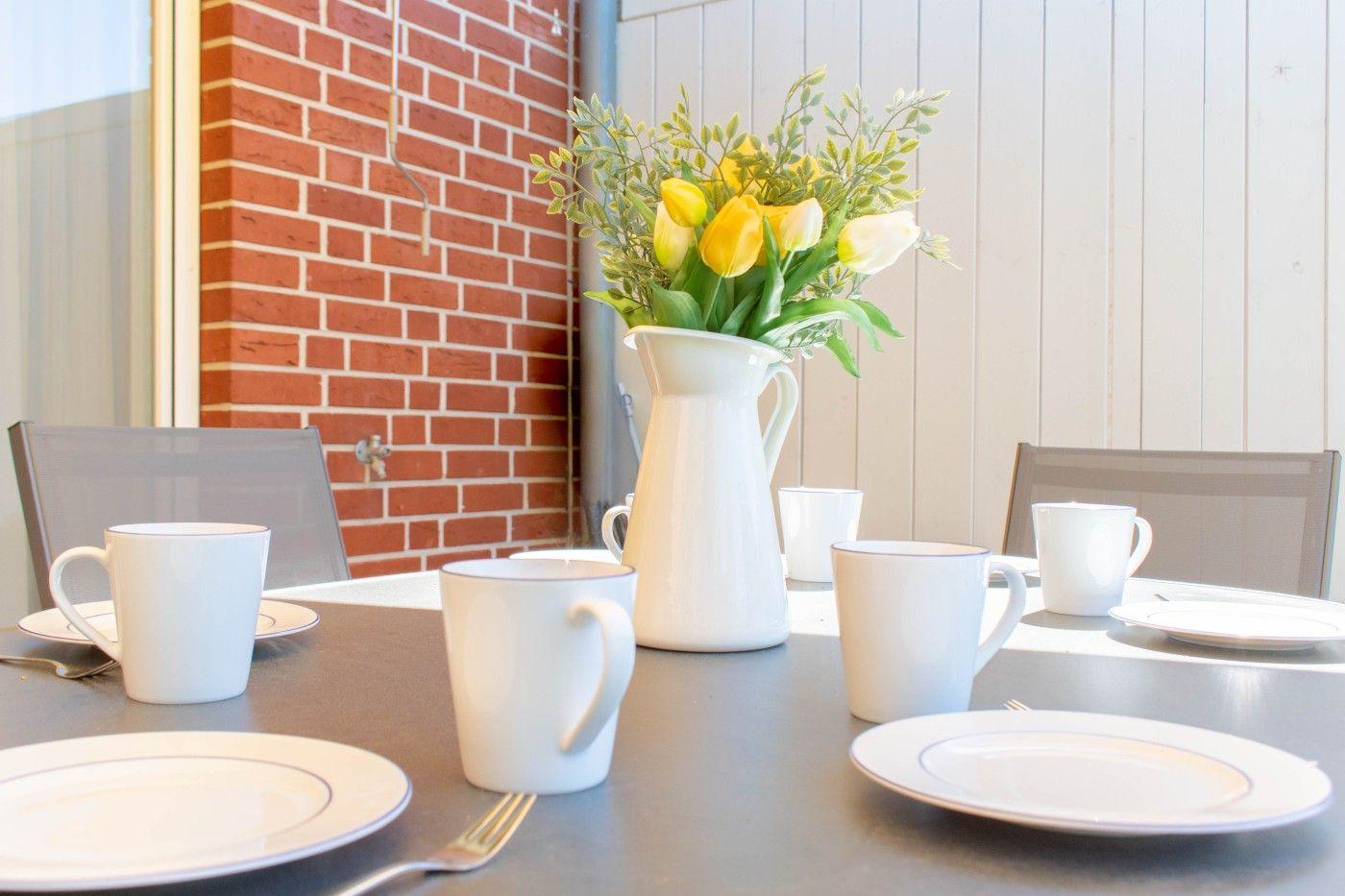 Tisch mit Tassen, Tellern und Blumen in weißem Krug vor rotem Ziegel- und weißem Holzwand.