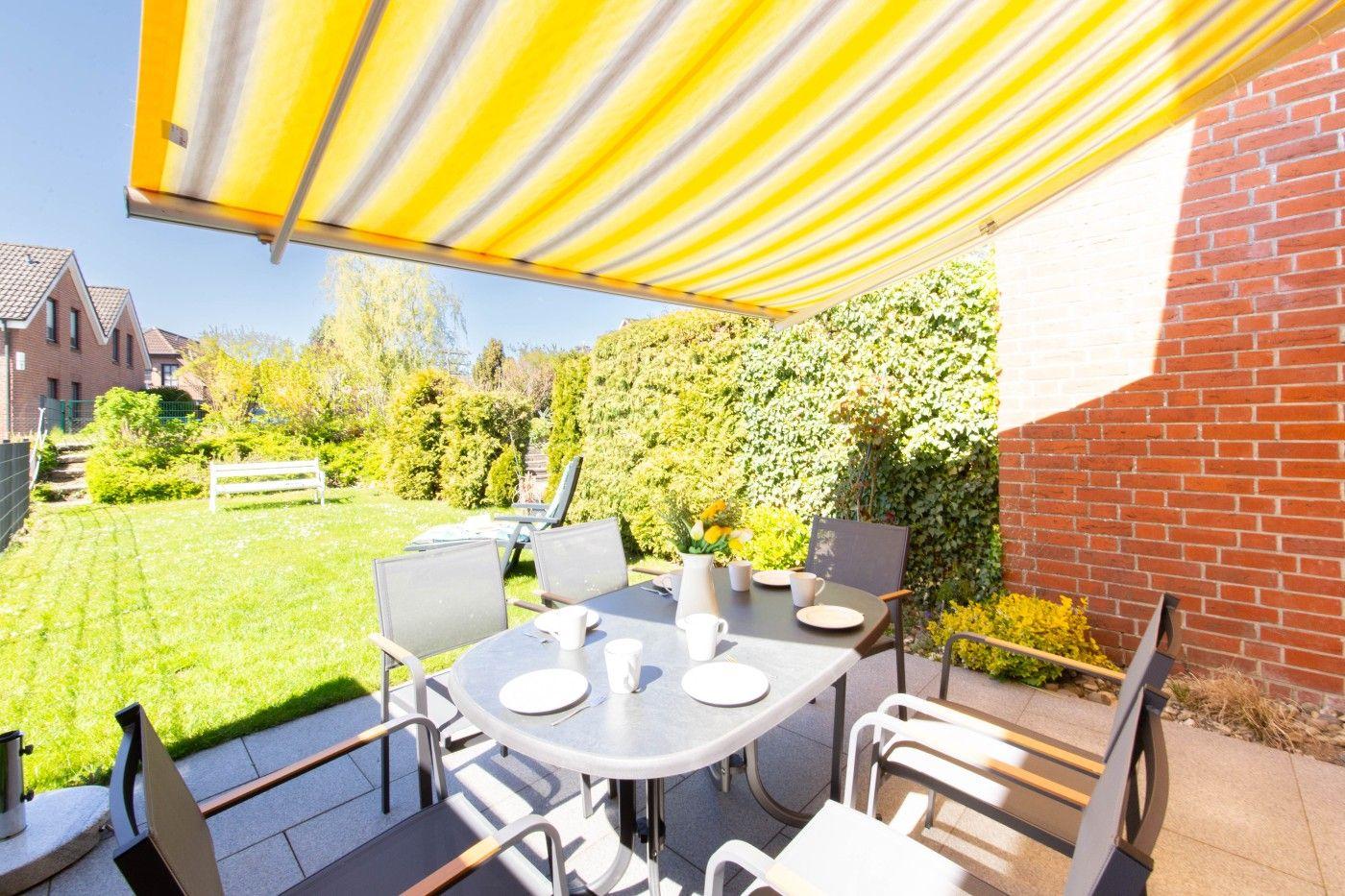 Terrasse mit Tisch, Stühlen und gelb-weißem Sonnenschutz. Grünflächen und Ziersträucher im Hintergrund.