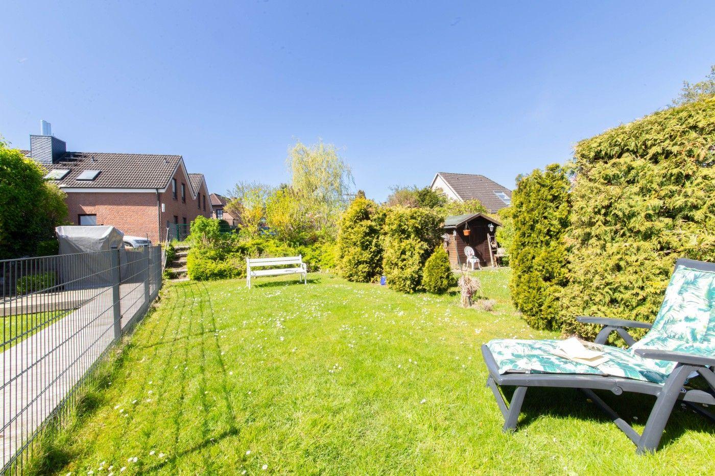 Garten mit Liegestuhl, Bank und Holzschuppen. Grasfläche, Hecken und Nachbarhäuser im Hintergrund.