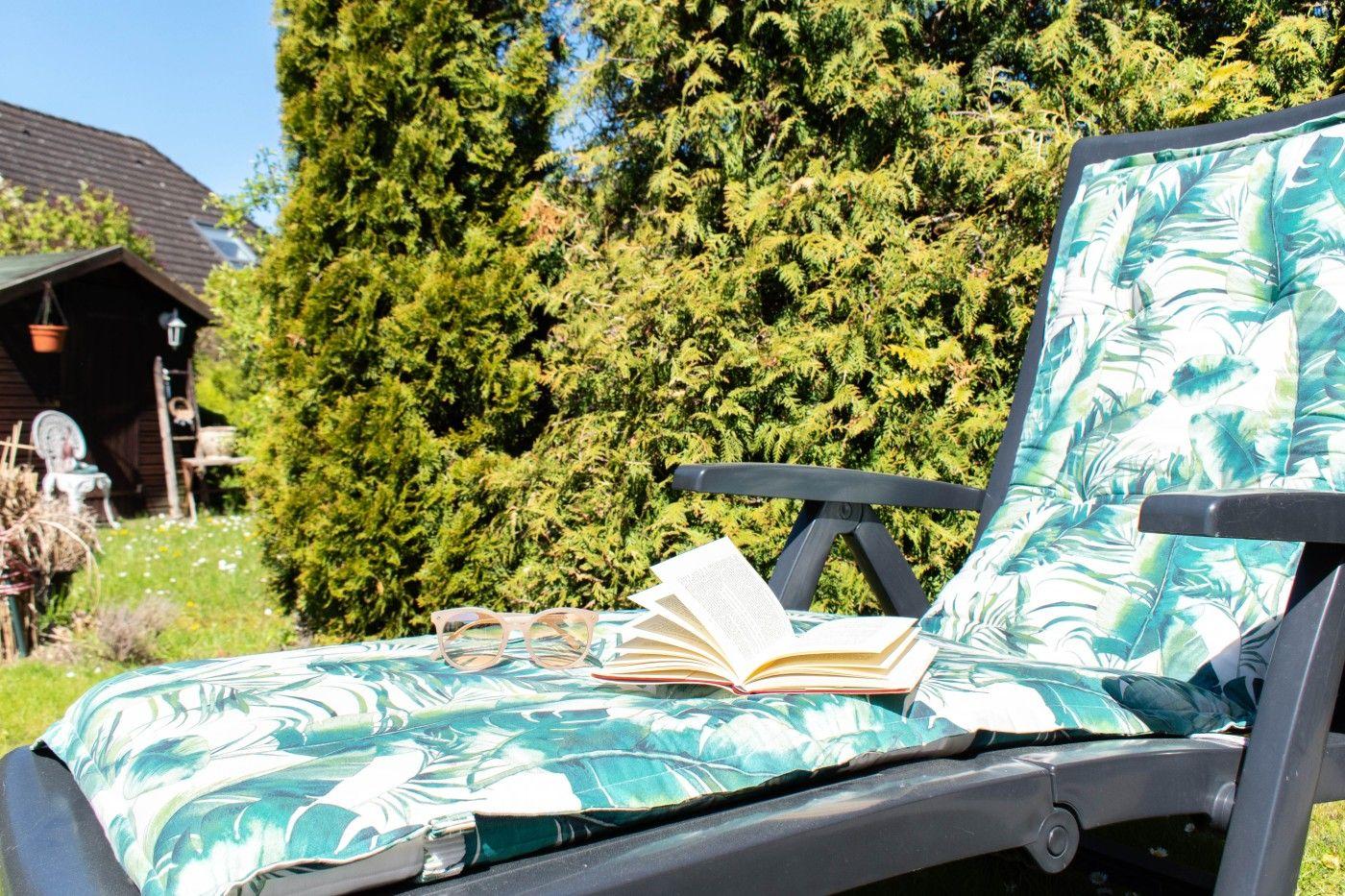 Liegestuhl mit Blattmuster, Buch und Brille im Garten vor grüner Hecke.