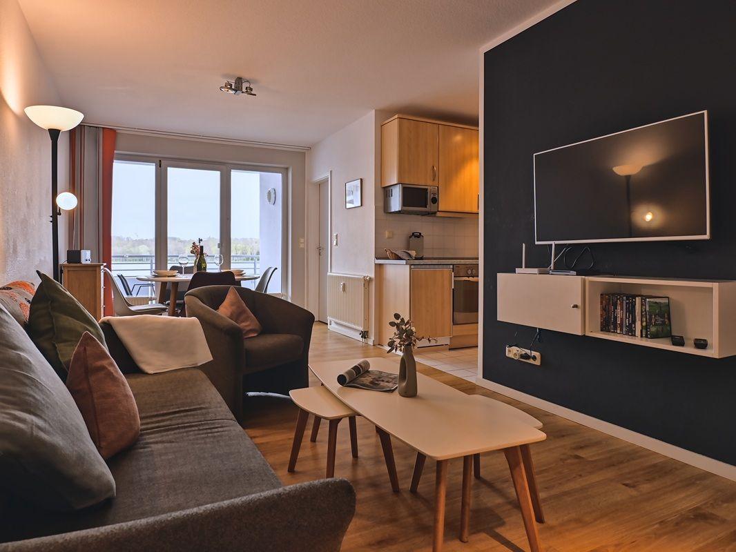 Wohnzimmer mit Sofa, Couchtisch und TV an schwarzer Wand. Küche und Balkon sind sichtbar.
