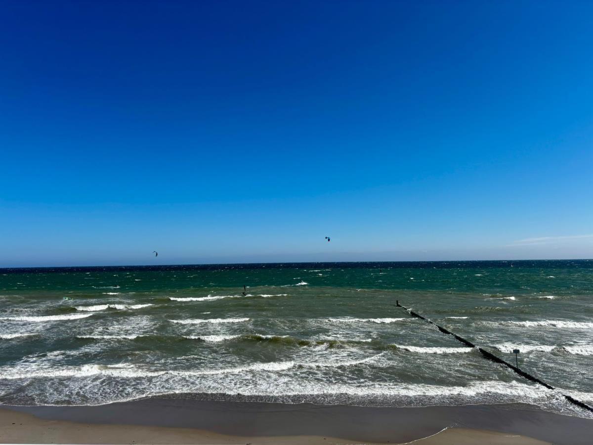 Strand mit Wellen und Kitesurfern unter blauem Himmel.