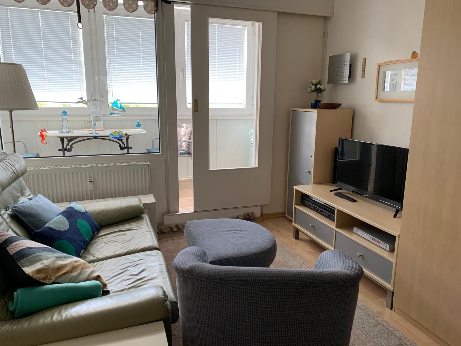 Wohnzimmer mit Sofa, Sessel und TV. Durchgang zur Terrasse mit Fenster.
