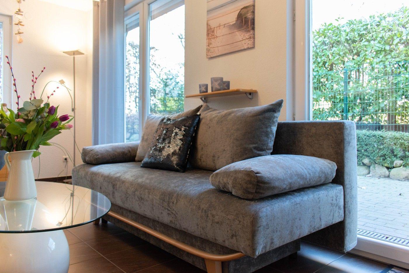 Ein grauer Sofa mit Kissen steht vor großen Fenstern. Auf einem Tisch steht eine Blumenvase.