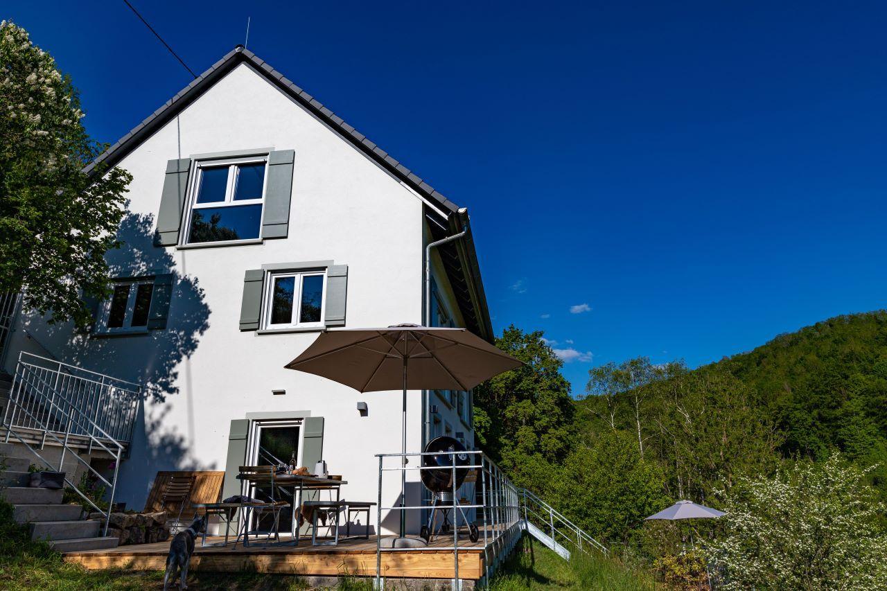 Wohnhaus mit Terrasse, Sonnenschirm und Grill. Umgeben von Wald und blauem Himmel.