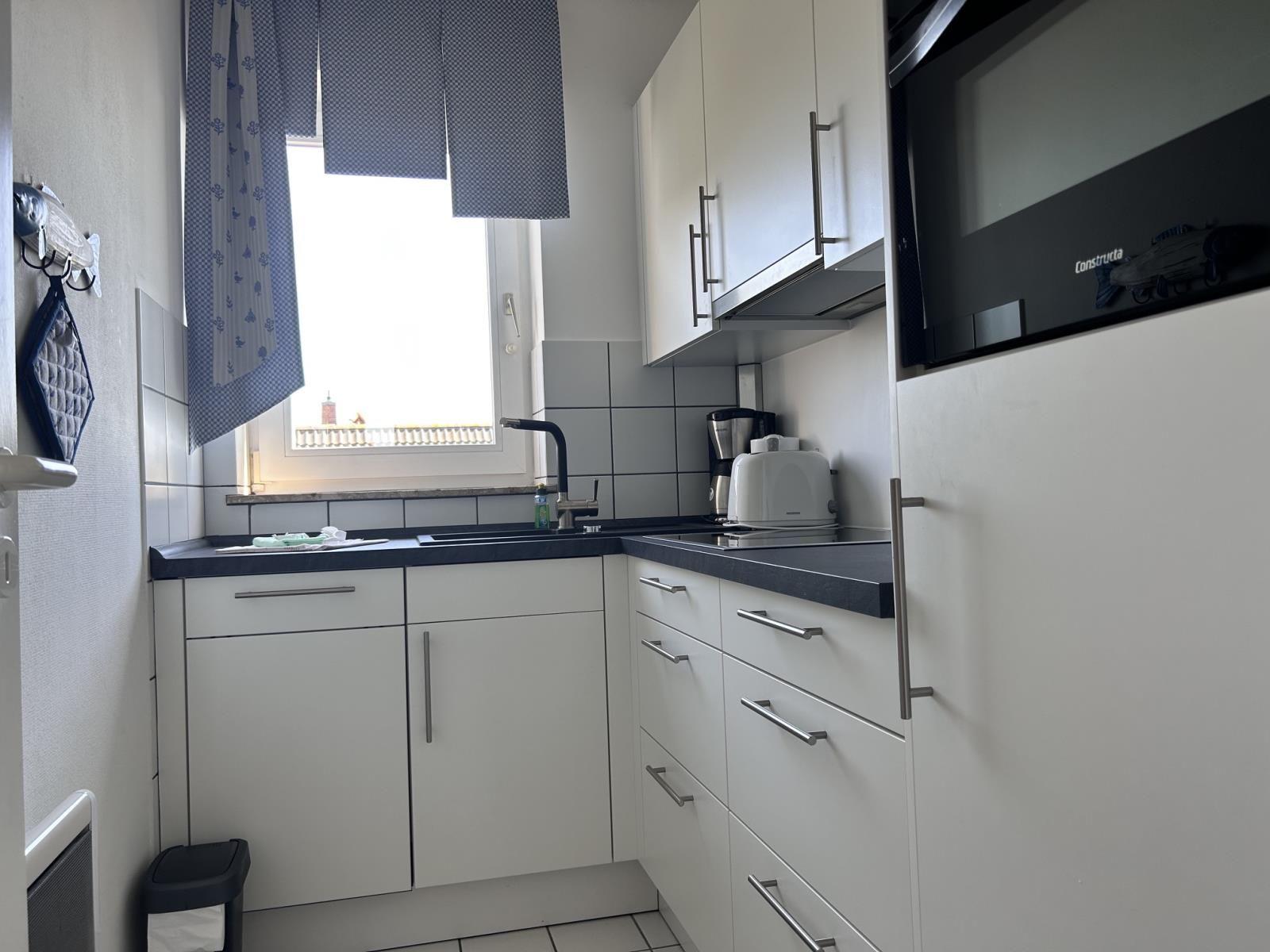 Küche mit weißen Schränken, dunkler Arbeitsplatte und Fenster mit Vorhängen.