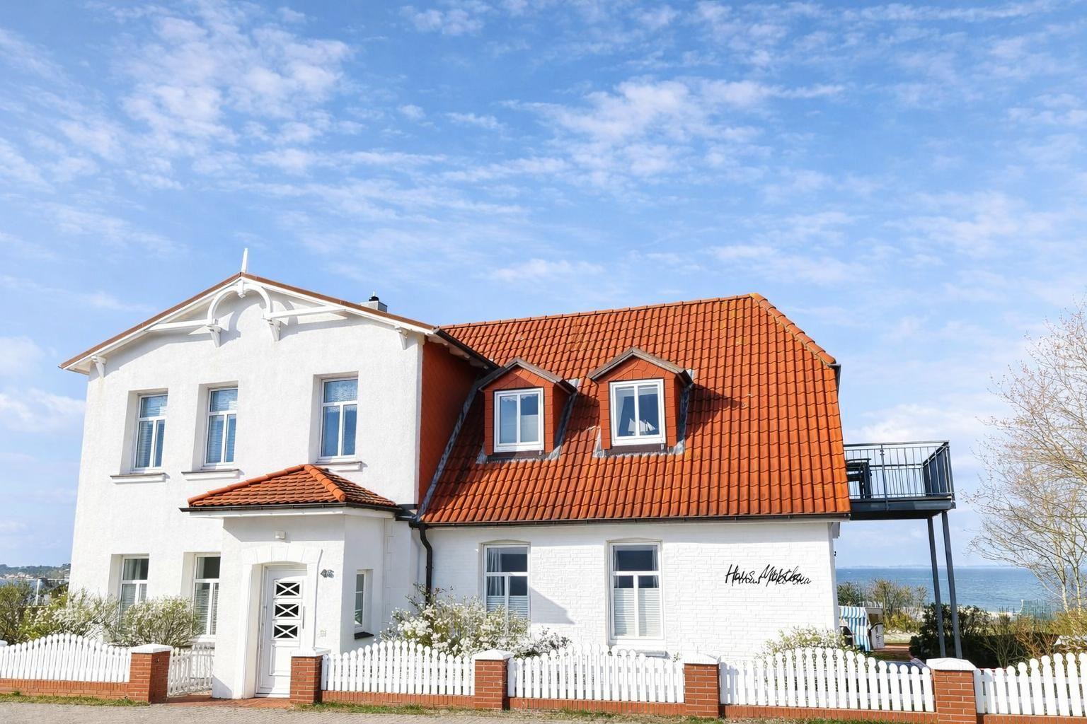 House with red roof and white facade, surrounded by a white fence with sea view.