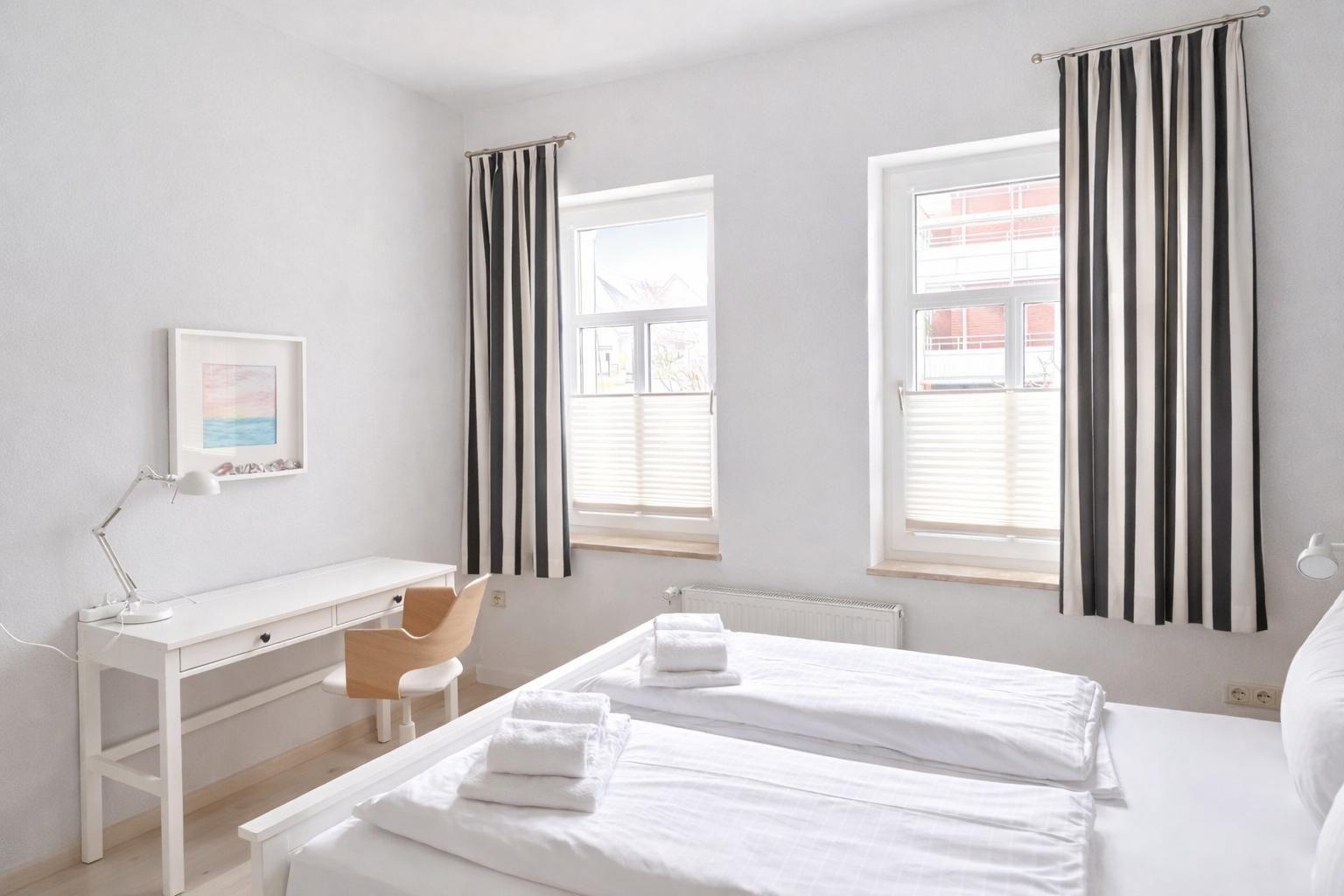Room with two beds, desk, and two windows with blinds and striped curtains.